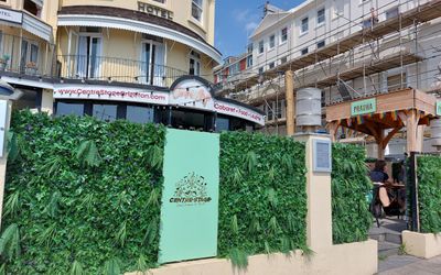 A wall of palnts with a sign reading Centre Stage with the laughing and crying drama masks on it, in the background there is a beer garden and a seafront regency building
