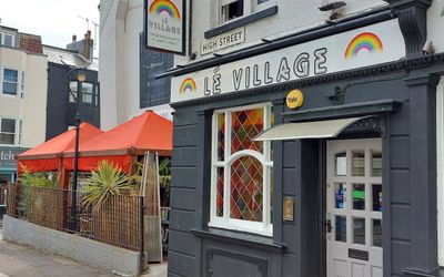 Side view of a grey pub called Le Village with a side garden with red umbrellas.