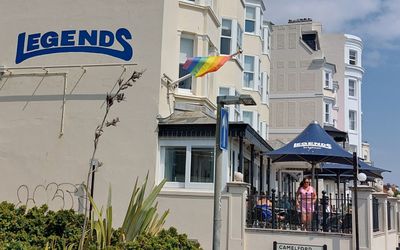 Side view of a seafront bar with terrace and blue parasols, on the side of the building is the bar name, Legends, and a rainbow Pride flag is flying.