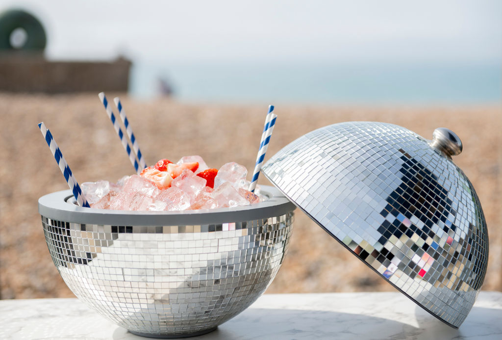 An ice bucket which resembles a disco ball is full of ice and cocktails with blue and white paper straws poking out, in the background you can see a pebbled beach and the sea