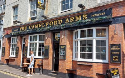 The exterior or a traditional looking pub with red brick and three bay windows and a sign in gold on black reading The Camelford Arms. Gay Bars Brighton Guide. LGBTQ+