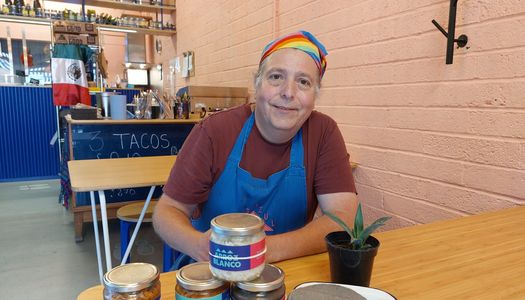 A man in a rainbow bandana and blue apron sits at a table with jars of food, tacos and a houseplant on it