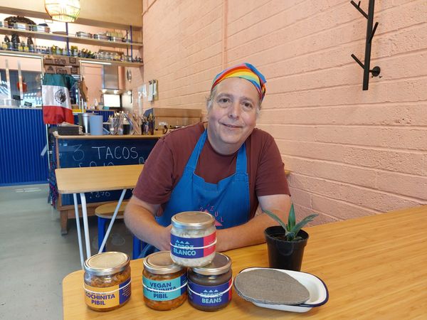 A man in a rainbow bandana and blue apron sits at a table with jars of food, tacos and a houseplant on it