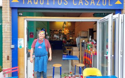 A man in a blue apron and rainbow bandanna stands outside a cafe called Taquitos Casazul. Mexican street food in Brighton