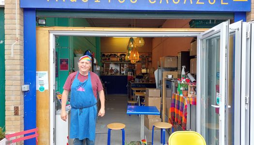 A man in a blue apron and rainbow bandanna stands outside a cafe called Taquitos Casazul