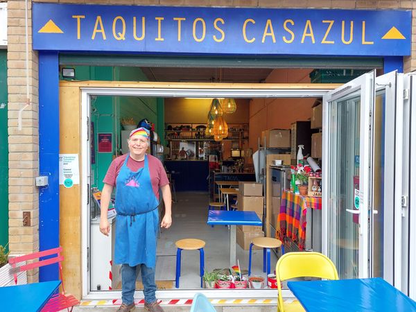 A man in a blue apron and rainbow bandanna stands outside a cafe called Taquitos Casazul