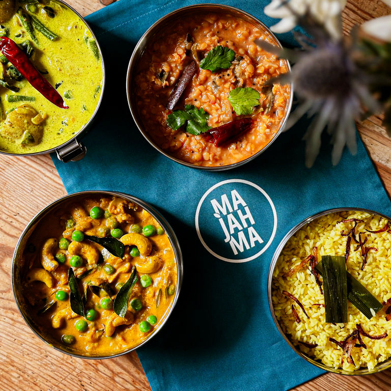 over head shot of four Majahma tiffins with yellow and orange Indian dishes, all served on dark blue Majahma cloth