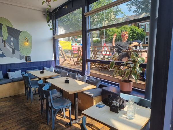 Inside a cafe, large windows let in light which spills over the tables, outside there is a terrace a view of trees