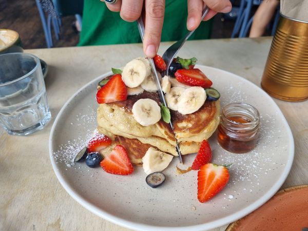 A stack of American pancakes covered in strawberries and bananas is being cut into.