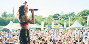 Mel C at Pub in the Park, dressed in jogging bottoms with a red, green and yellow stripe and a sports top in red, black and blue is on stage singing into a microphone, in the background is a crowd of spectators in a sunny park with tents in the background