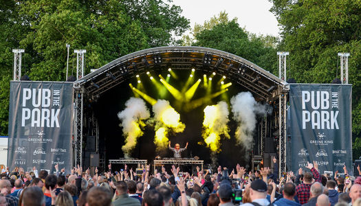 Things to do in Brighton this summer. Main stage at pub in the park, a crowd stand before the domes stage while a DJ performs and clouds of smoke and lime green lights flash. In the background trees tower over the stage and a clear sky shines through