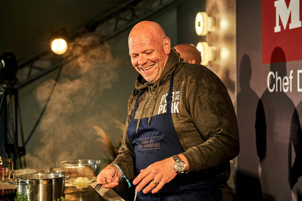 Chef Tom Kerridge in a green hoodie and blue apron on a stage, doing a food demonstration