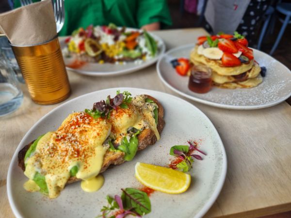 three plates of brunch, nearest is a Benedict on toast, behind that a plate of American pancakes and beyond that a colourful smash of summer vegetables on sourdough