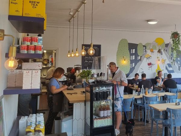 A busy cafe, with diners sat at blue charis, a wall is painted with a mural of dancing ingredients playing insturments, a man with a dog makes a purchase at the counter