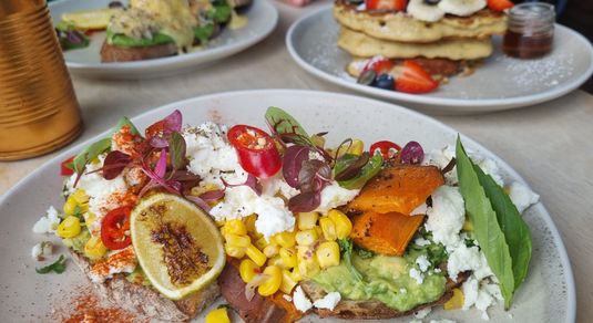 A colourful plate of avocado on toast with sweetcorn, sweet potato, feta and chillis, behind is another plate of American pancakes with berries and banana and another with an egg Benedict