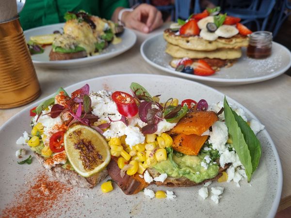 A colourful plate of avocado on toast with sweetcorn, sweet potato, feta and chillis, behind is another plate of American pancakes with berries and banana and another with an egg Benedict