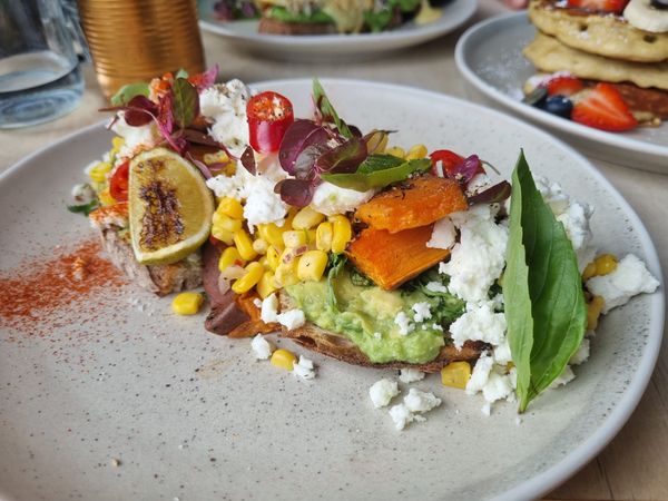 A colourful brunch of avocado on sourdough topped with a mix of bright yellow sweetcorn, sweet potato, feta, fresh red chilis and burnt lime wedges