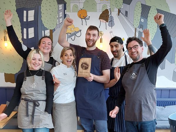 Six workers in a cafe stand together arms upraised in celebration, holding a wooden trophy, behind them is a mural of ingredients dancing and playing musical instruments walking down a residential street. Queens Park cafe