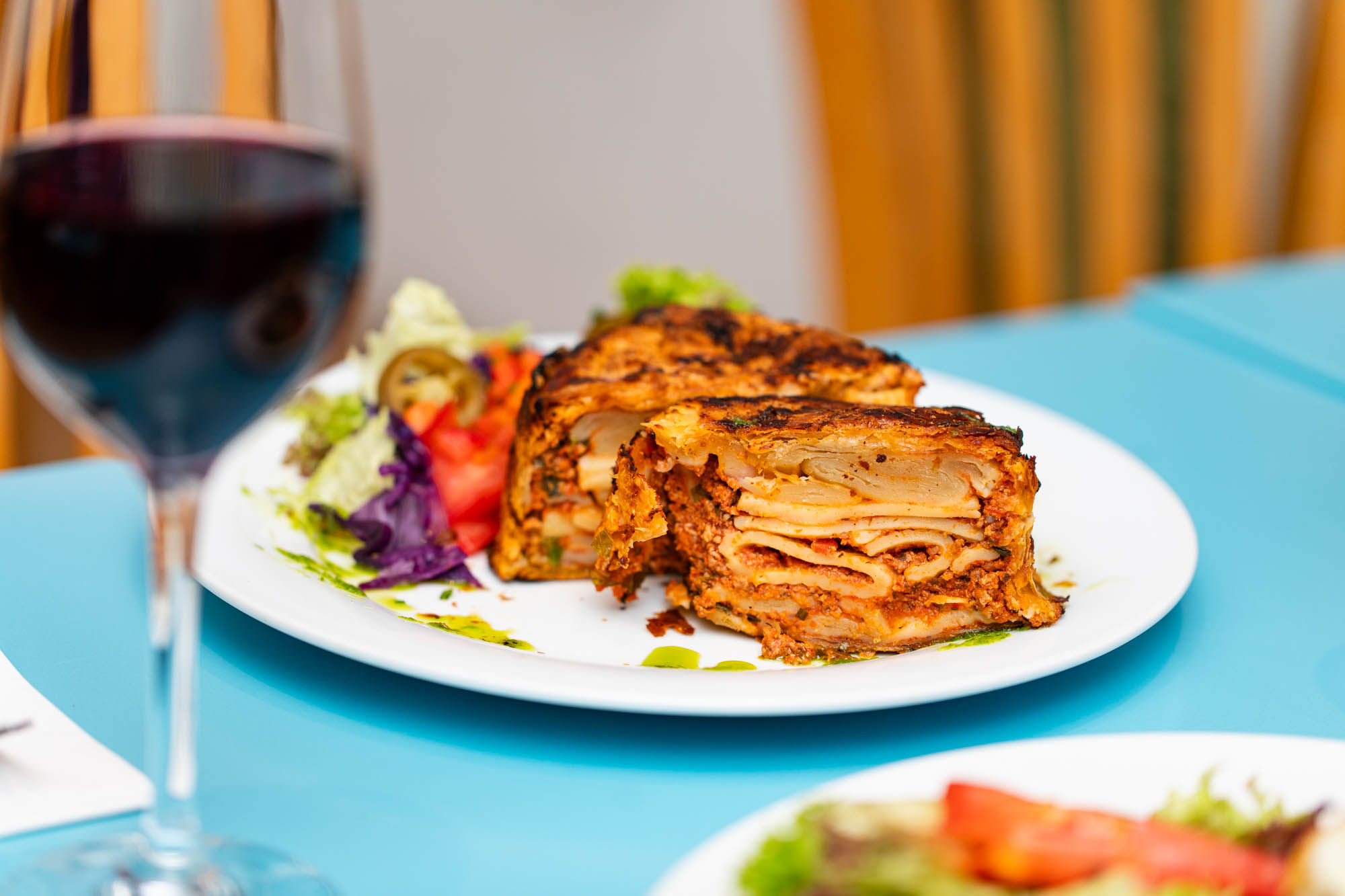 A plate of lasagne and salad with a glass of red wine served on a blue table.