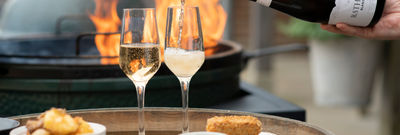 Two flutes are being filled with sparkling wine from a bottle of Rathfinny Estate Blank de Noirs, they're on the top of a wine barrel with two plates of food, one of potatoes the other croquettes behind them a grill flames up. Available for Alcohol Delivery Brighton
