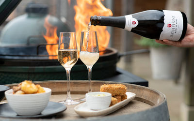 Two flutes are being filled with sparkling wine from a bottle of Rathfinny Estate Blank de Noirs, they're on the top of a wine barrel with two plates of food, one of potatoes the other croquettes behind them a grill flames up. Available for Alcohol Delivery Brighton