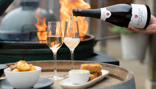 Two flutes are being filled with sparkling wine from a bottle of Rathfinny Estate Blank de Noirs, they're on the top of a wine barrel with two plates of food, one of potatoes the other croquettes behind them a grill flames up. Available for Alcohol Delivery Brighton