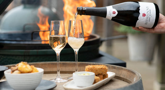 Two flutes are being filled with sparkling wine from a bottle of Rathfinny Estate Blank de Noirs, they're on the top of a wine barrel with two plates of food, one of potatoes the other croquettes behind them a grill flames up. Available for Alcohol Delivery Brighton