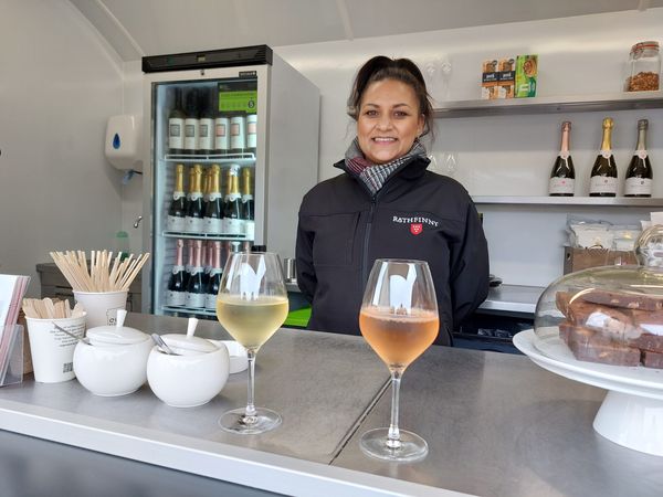 A woman in a mobile catering van stands in front of a glass of white and rosé wine, in the background there's a fridge full of wine bottles