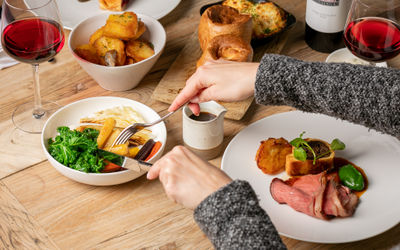 A wooden table is laid with the elements of a Sunday lunch, a plate of beef with gravy, a board with Yorkshire puddings and a skillet of cauliflower cheese, a deep bowl of roast potatoes and another of roasted rood vegetables and kale, two glasses of cherry red wine are on the table and arms in a grey jumper reach over to the bowl of veg with cutlery