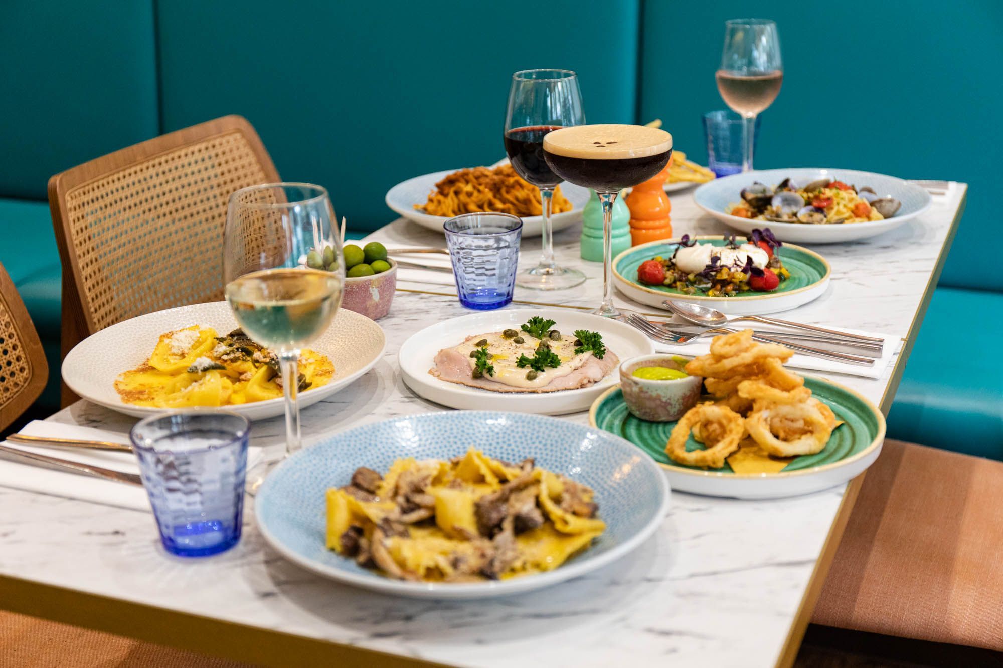 side shot of the white table laid out with delicious Italian dishes inluding different pastas, served with glasses of wine and cocktails