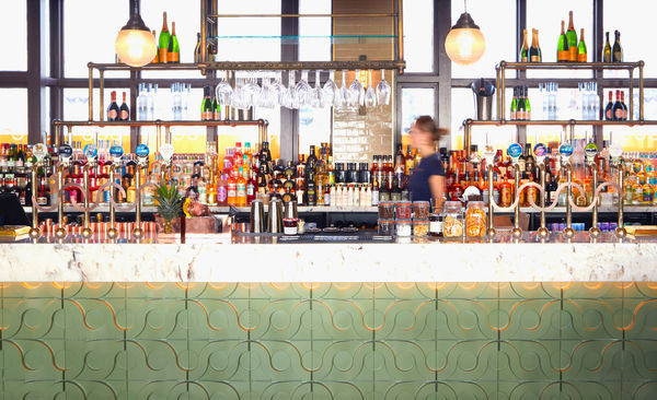 A picture of a modern bar with green tiles and silver pumps. A waiter passes through the image. 