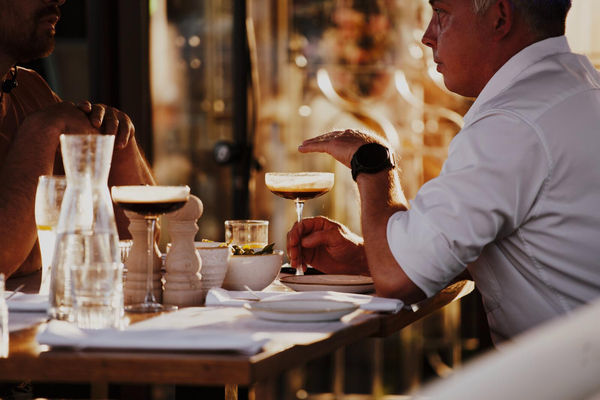 A man and woman sitting at a dining table with elegant cocktails and a carafe of water. They are speaking. 