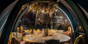 Inside an igloo pod, a round wooden table with dark gold coloured tea light holders, chairs and a comfy cushioned bench surround the table and are draped with blankets. In the top of the igloo hands a display of dried flowers, grasses and lights. This picture represents the start of the Brighton igloo guide and it is popular in many Brighton restaurants.