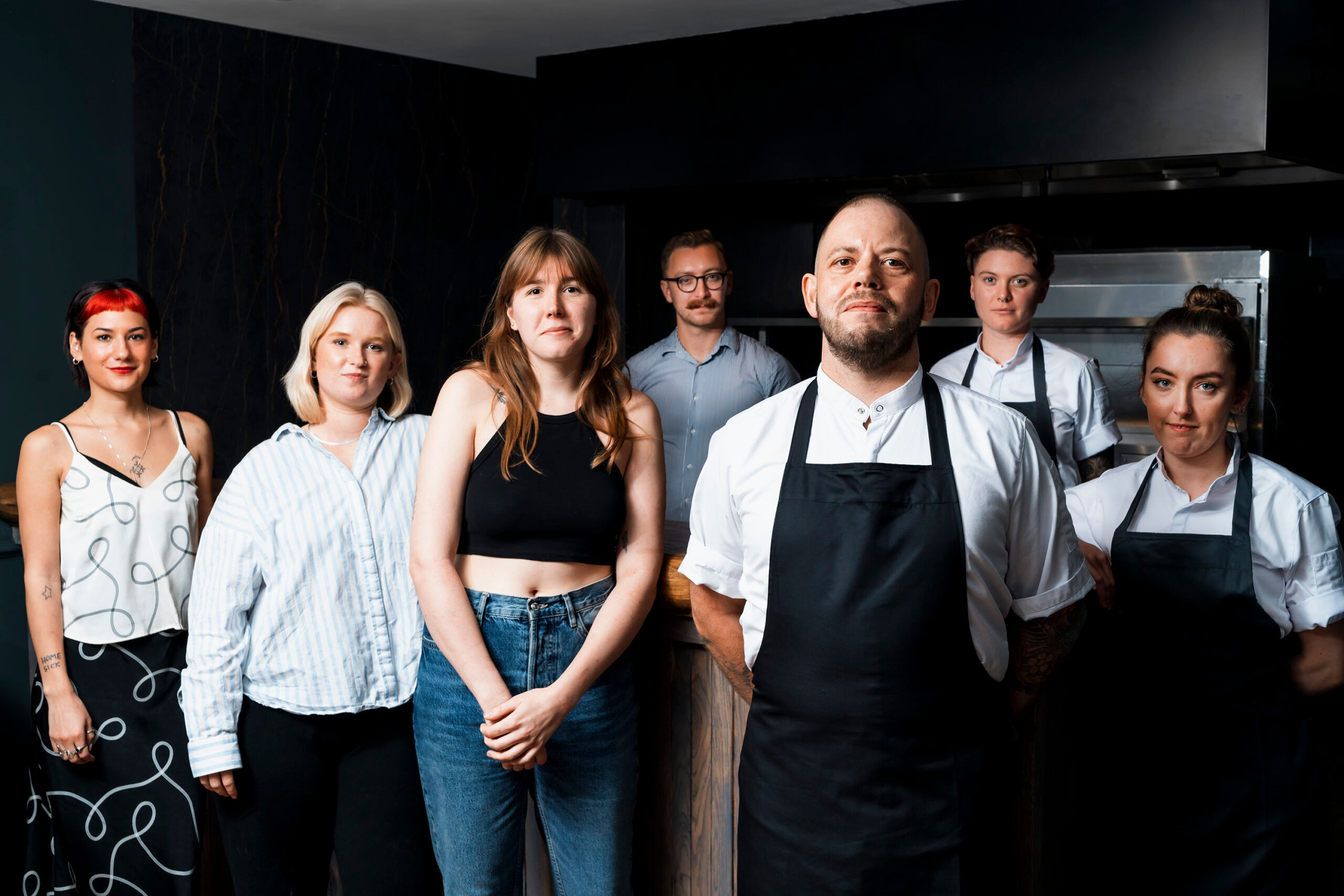 The new team at Furna, a group of seven people stand in a darkened room, at the front is a bald man in chef whites and a navy apron, to his left a female chef dressed the same