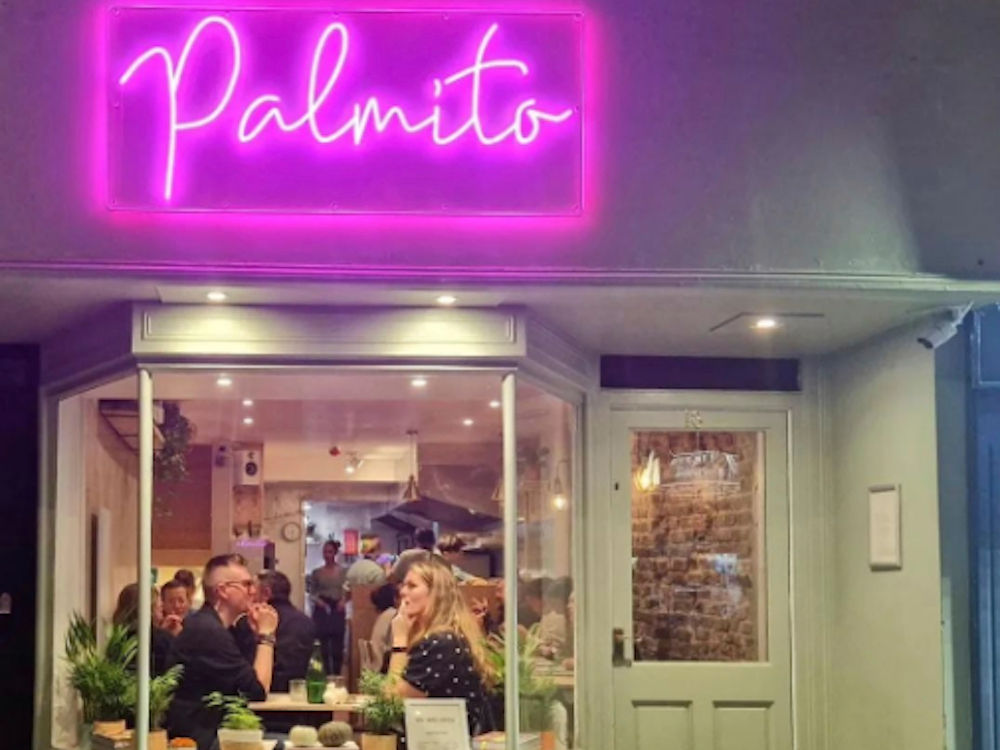 Exterior of a restaurant with sage green exterior, a large window shoes diners sat at tables behind them are brick walls. Over the window is a large neon sign saying Palmito