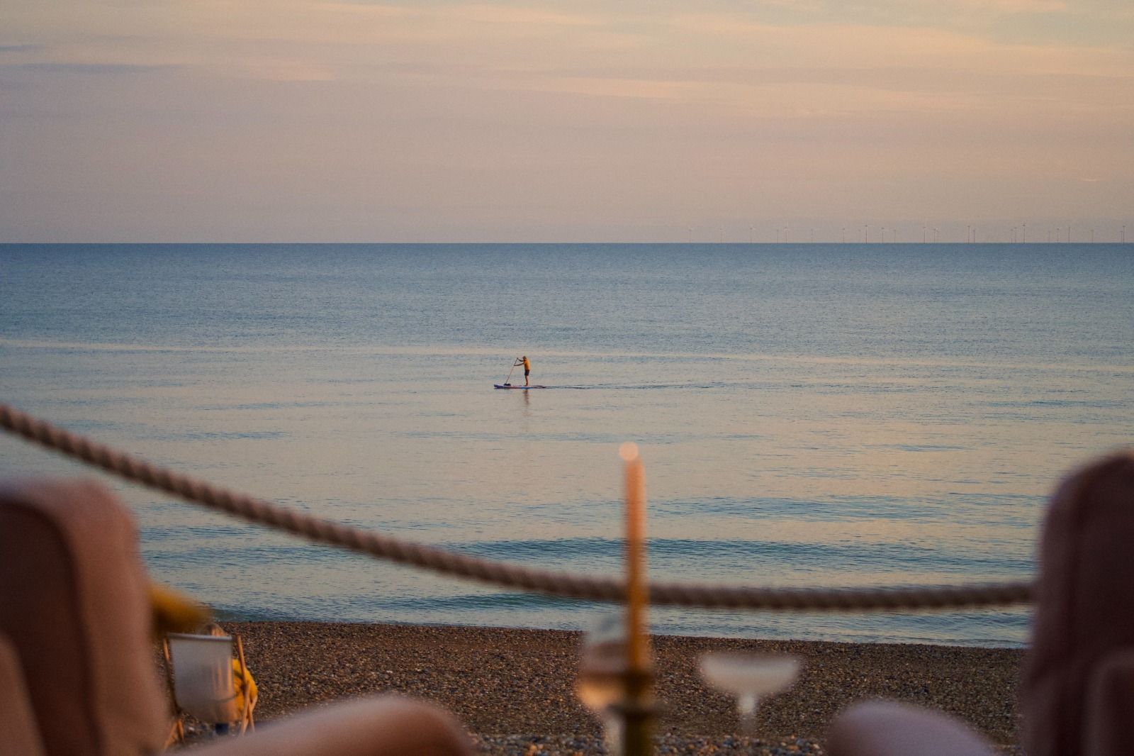 A view over a calm sea at sunset, in the foreground just out of focus are two chairs, a glass of wine, a cocktails and a single candle