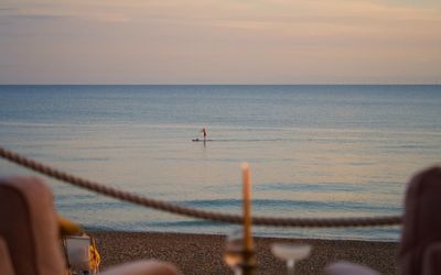 A view over a calm sea at sunset, in the foreground just out of focus are two chairs, a glass of wine, a cocktails and a single candle
