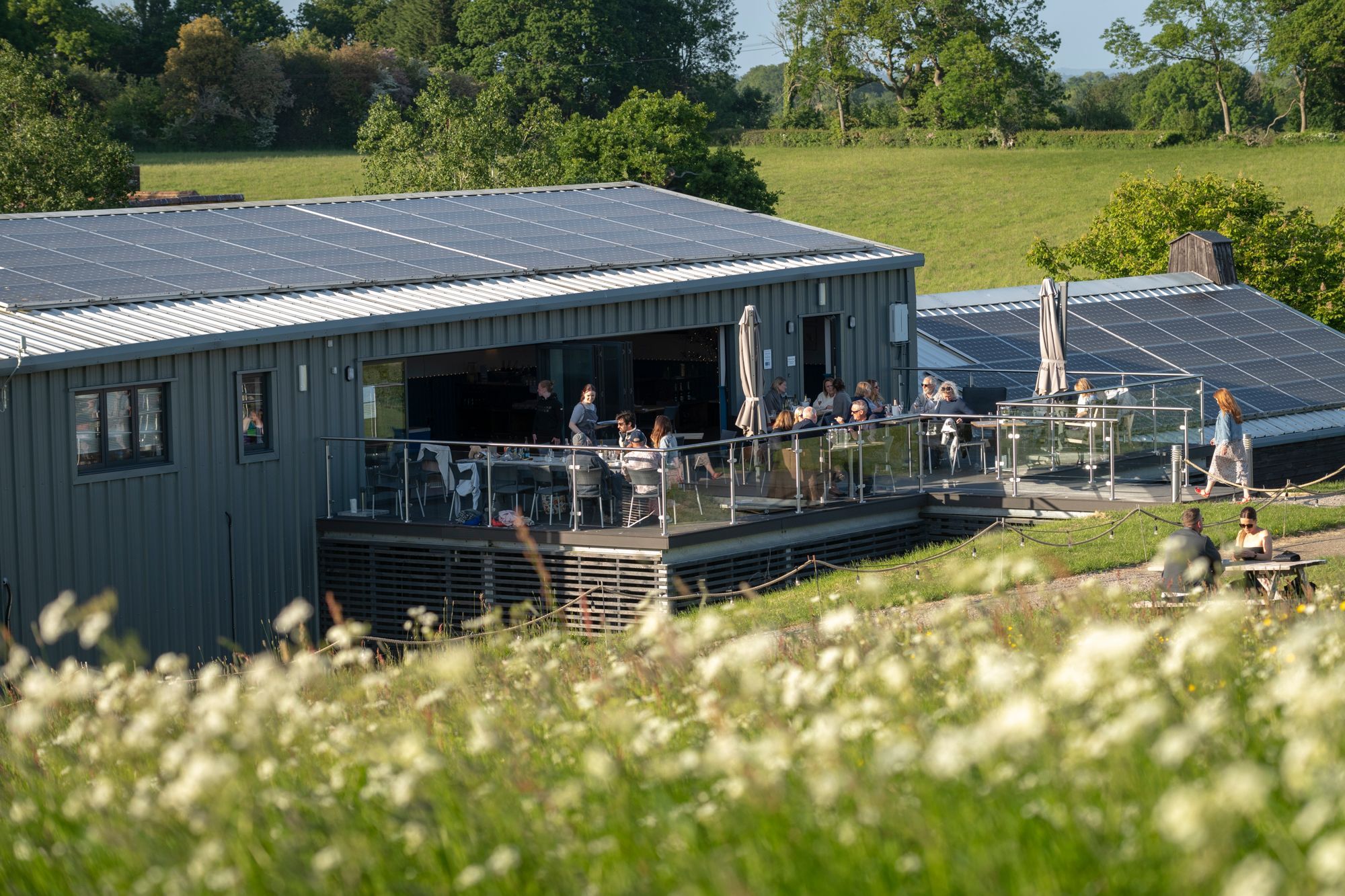 shot of the Albourne Estate from the distance, grey building with the solar panels on the roof and people sitting at the terrace