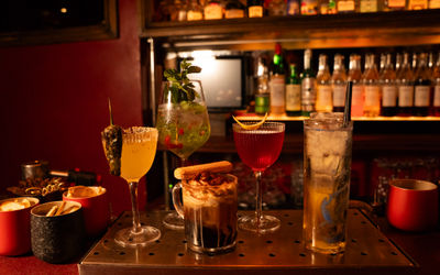 A richly atmospheric lounge scene in deep red tones. In the foreground, a tufted velvet banquette lines one wall, beside small round marble or dark wood tables and low stools. On the wall above, framed vintage photographs or art pieces are arranged in a gallery-style layout. To one side, a wall lamp with a fringed shade glows warmly, casting soft light across the seating. In the background, shelves of spirits and glassware peek through, hinting at a bar behind a semi-obscured partition. The mood is intimate, plush, and evocative of a 1920s speakeasy with modern elegance.