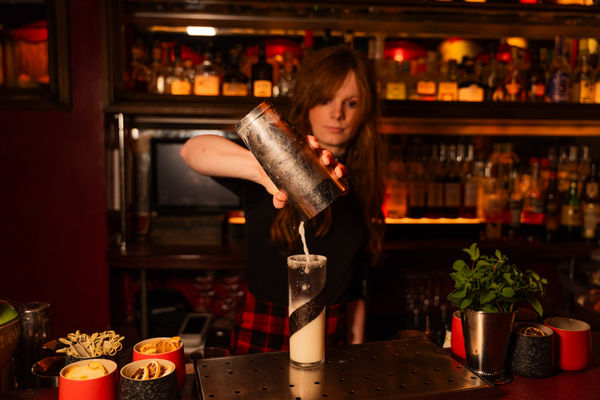 A moody, intimate cocktail bar interior bathed in warm red light. Plush velvet seating lines the walls, with framed artwork and vintage-style décor displayed above. A low-slung bar counter stretches into the scene, topped with bottles, glassware, and ambient lighting. Soft, fringed lampshades cast a golden glow across the space, accentuating rich tones and casting gentle shadows.