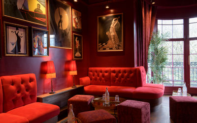 A wide-angle view of a stylish speakeasy-style cocktail lounge with rich red lighting and plush, dark furnishings. In the foreground, low tables and deep velvet sofas invite relaxed seating. Along one side, a polished bar counter runs with display shelves of bottles and soft, hanging fringed lamps casting a warm amber glow. On the opposite side, tufted bench seating spans beneath walls adorned with framed vintage artwork and mirrors. In the background, a large bay window opens onto a terrace or balcony area overlooking leafy treetops and glimpses of ornate architecture beyond. The overall mood is intimate, retro-glamorous, and inviting for a sophisticated evening out.