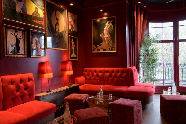 A wide-angle view of a stylish speakeasy-style cocktail lounge with rich red lighting and plush, dark furnishings. In the foreground, low tables and deep velvet sofas invite relaxed seating. Along one side, a polished bar counter runs with display shelves of bottles and soft, hanging fringed lamps casting a warm amber glow. On the opposite side, tufted bench seating spans beneath walls adorned with framed vintage artwork and mirrors. In the background, a large bay window opens onto a terrace or balcony area overlooking leafy treetops and glimpses of ornate architecture beyond. The overall mood is intimate, retro-glamorous, and inviting for a sophisticated evening out.