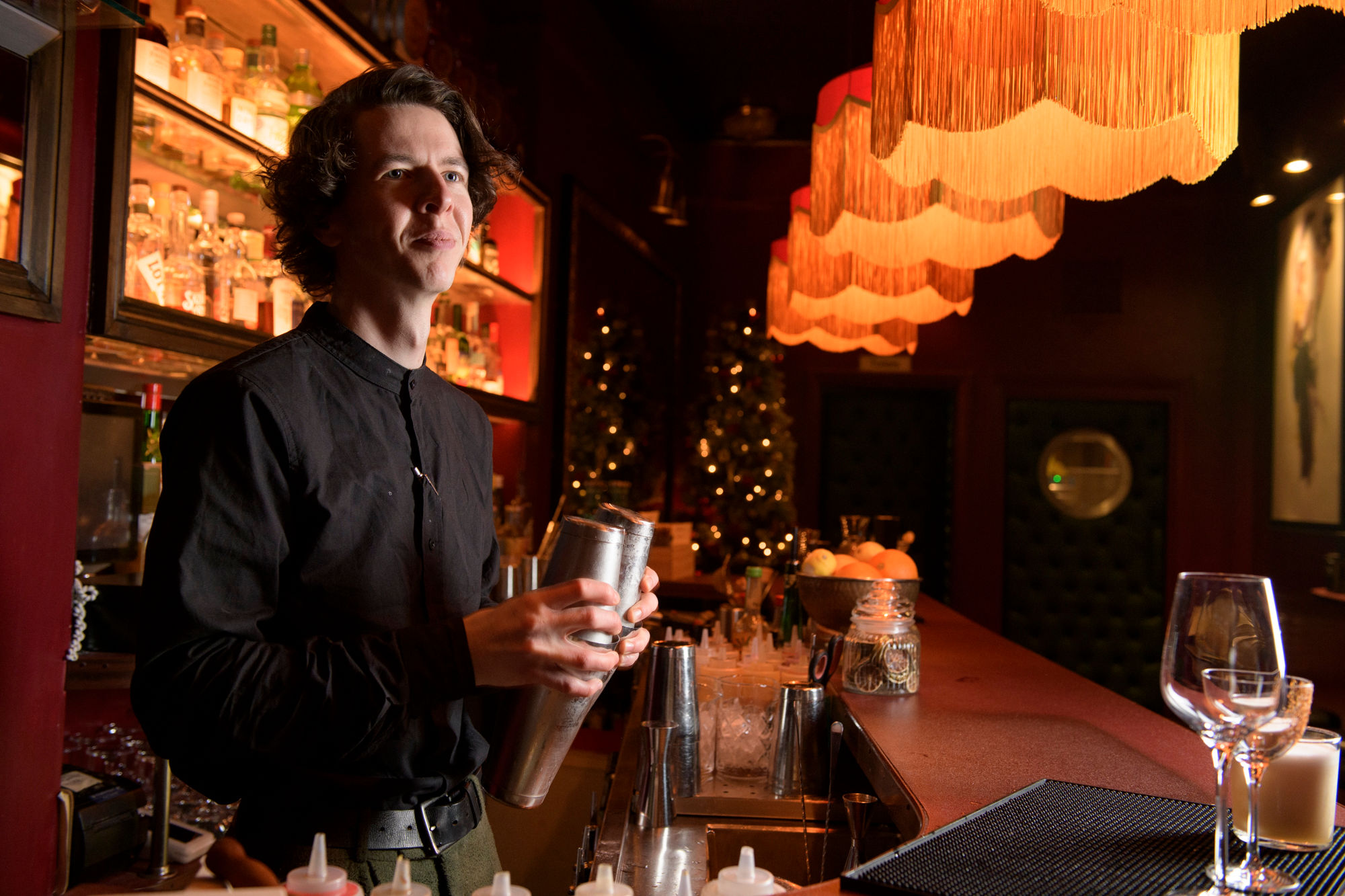 A man with long hair wearing a dark collarless shirt stands behind a bar holding a cocktail shaker, a row of tasseled lamps stretch off into the distance over the bar, into a dark background