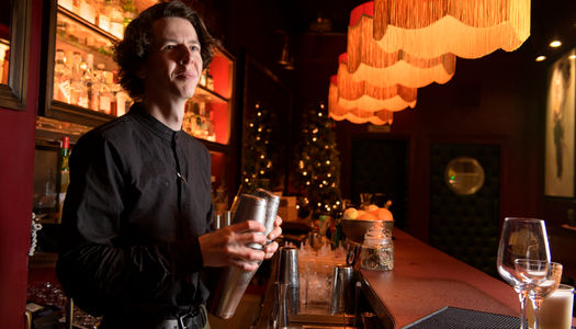 A man with long hair wearing a dark collarless shirt stands behind a bar holding a cocktail shaker, a row of tasseled lamps stretch off into the distance over the bar, into a dark background - Bar Valentino Brighton. Alcohol free Brighton