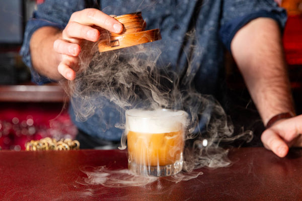 an amber cocktail has smoke coming from the top of it. A man is pulling the lid off to release smoke from the glass.