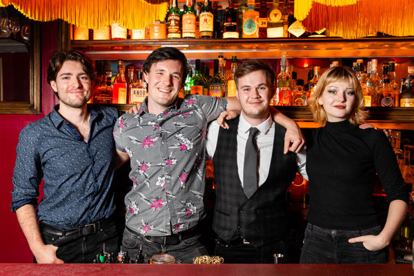 4 adult people who work at a bar behind the bar hugging. 3 white men and one white lady. 
