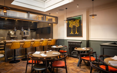Inside Furna restaurant Brighton, arow of mustard padded bar seats line the open plan kitchen, the main restaurant has a lattice parquet floor and round wooden tables, an iron work fireplace sits in a green panelled wall and artwork depicts stylised trees