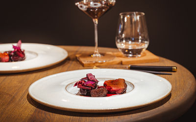 A dish at Furna Restaurant Brighton. Salt aged Sika deer on a deep rimmed flat plate on a soft edged wooden table, a glass of wine and water are in the background