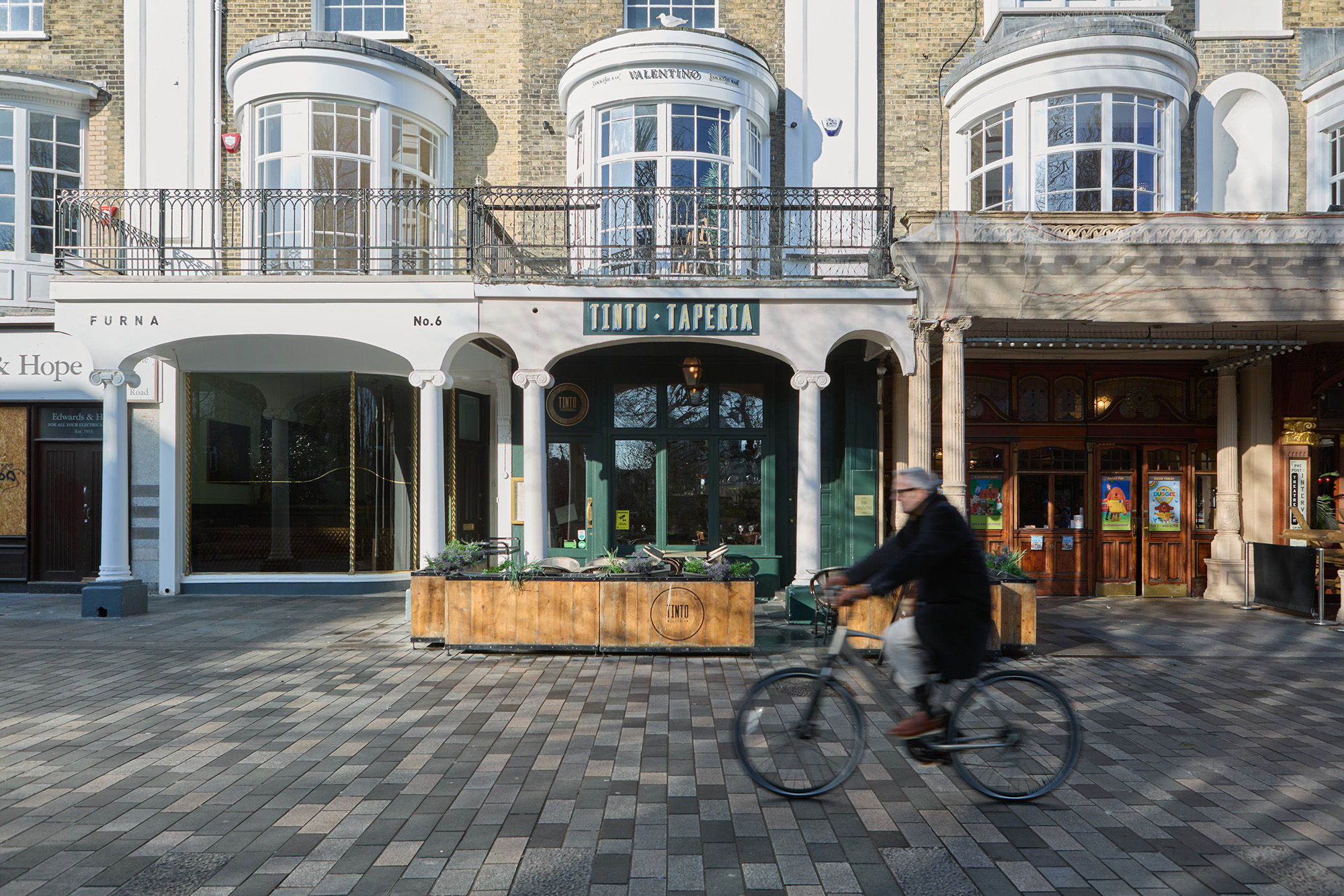 A wide exterior photograph of Tinto Taperia Brighton with a cyclist out of focus riding past the building along the path. There's green paintwork on the building and a flower bed around the terrace.
