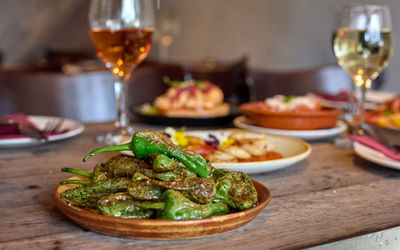 A pile of padron peppers served on a brown plate with a glass of wine in the background. Lots of other plates of food in the background on a wooden table at Tinto Taperia Brighton.